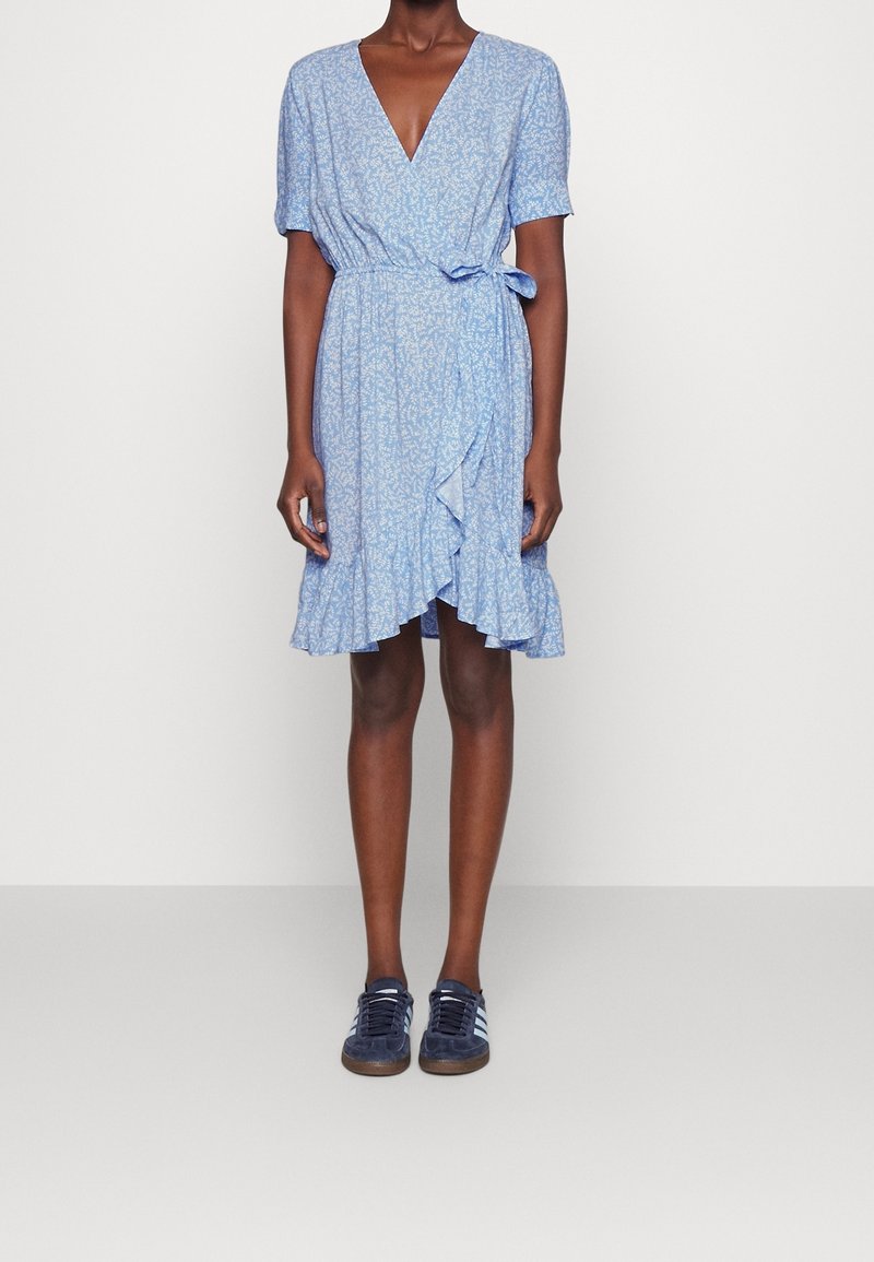 Personne portant une robe portefeuille fleurie bleu clair longueur genou, avec ourlet à volants et baskets bleu marine sur un fond uni clair.