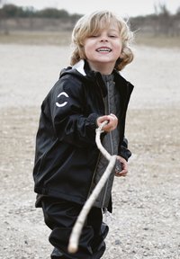 Črna vodoodporna jakna z belimi poudarki, sivim odebeljenim notranjim slojem in elastičnimi manšetami. Otrok drži palico, se smeji, stoji na prodnatem površju.