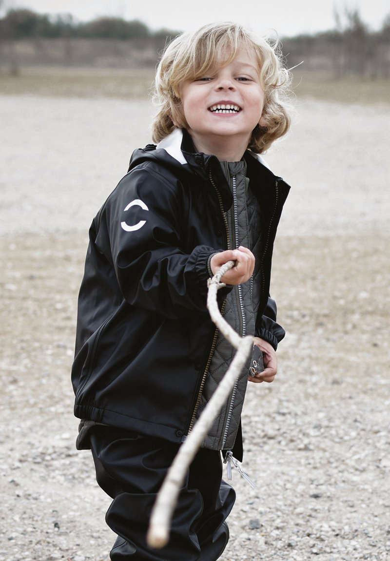Črna vodoodporna jakna z belimi poudarki, sivim odebeljenim notranjim slojem in elastičnimi manšetami. Otrok drži palico, se smeji, stoji na prodnatem površju.