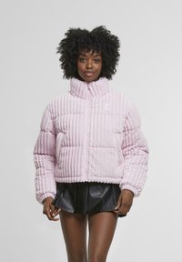 Giacca trapuntata rosa con texture a coste verticali, collo alto, chiusura con zip e logo ricamato sul petto. Abbinata a una gonna nera.