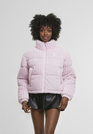 Giacca trapuntata rosa con texture a coste verticali, collo alto, chiusura con zip e logo ricamato sul petto. Abbinata a una gonna nera.