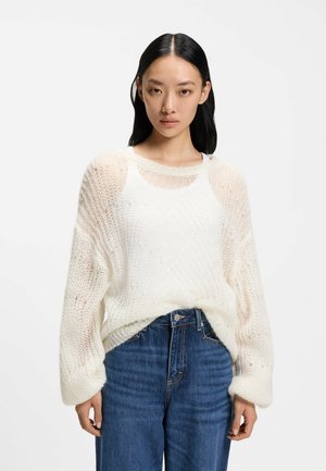 Junge Frau mit glatten schwarzen Haaren, die einen lockeren, weißen Strickpullover über einem weißen Oberteil und blaue hoch taillierte Jeans vor neutralem Hintergrund trägt.