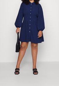 Sininen ruudullinen mekko, jossa on pitkät puffi-hihat, napitettava etuosa ja kaulus. Parita mustan olkalaukun ja litteiden mustien sandaalien kanssa.