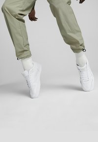 White athletic sneakers with perforated detailing and round laces, worn with light-colored joggers and crew-length socks against a gray background.