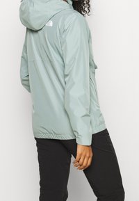 Light green, waterproof jacket with a hood. Features elastic cuffs and a loose fit. Logo on upper back. Worn over black pants.