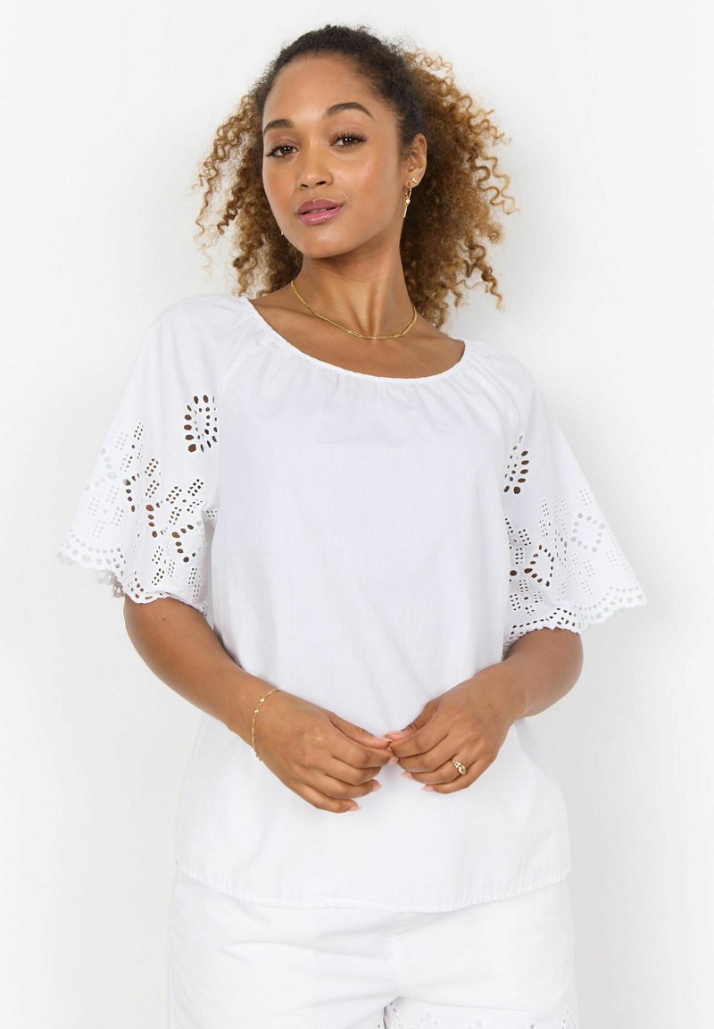 Blusa di cotone bianca con maniche corte ricamate. Presenta un colletto rotondo e una vestibilità morbida. Motivi intagliati visibili sui bordi delle maniche.