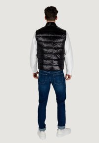 Chaleco de plumas negro con acabado brillante y secciones acolchadas amplias, usado sobre un sudadera blanca, combinado con vaqueros azules y zapatillas blancas.