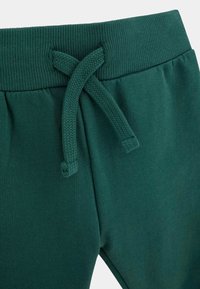 Žali medvilniniai džinsai turi išorinio audinio juosmenį su nertomis juostelėmis, surištomis į mazgą, išryškindami lygią tekstūrą ir elastinį pritaikymą.