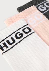 Trijų kojinių rinkinys: balta, rožinė ir tamsiai pilka, su gofruota tekstūra ir drąsiu juodu siuvinėtu "HUGO" logotipu.