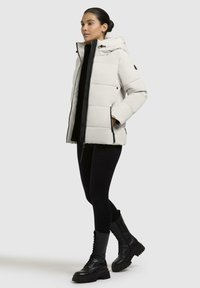 Giacca puffer off-white con zip nera sul davanti, cappuccio e tasche laterali. Caratterizzata da un design a quadrati trapuntati e polsini elastici. Indossata sopra leggings neri.