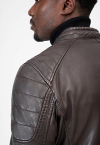 Braune Lederjacke mit gesteppten Schulterdetails und einem schwarzen, gerippten Kragen, die eine glatte Textur und ein elegantes Design zeigt.
