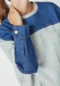 Denim and grey sweatshirt featuring a color-block design with a blue denim upper part and grey lower portion, accented by gold hardware on cuffs.