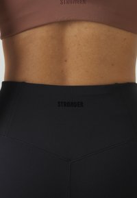 Crne tajice visokog struka s teksturiranom završnom obradom i crnim logom "STRONGER" na struku, u kombinaciji s smeškastim sportskim grudnjakom.