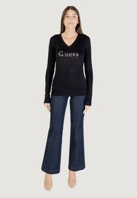 Schwarzer V-Ausschnitt-Pullover mit Strass "Guess"-Logo auf der Brust, getragen mit dunklen Jeans im Schlag und beigen Absätzen, der einen taillierten Look präsentiert.