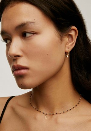 Joyería en tono dorado que incluye un collar tipo choker con cuentas y un pequeño colgante, junto con aretes de aro a juego con acentos decorativos. Textura de piel suave.