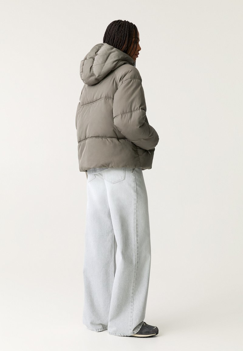 Giacca imbottita verde oliva con cappuccio, caratterizzata da un design trapuntato, indossata con jeans a gamba larga grigio chiaro e sneakers nere.