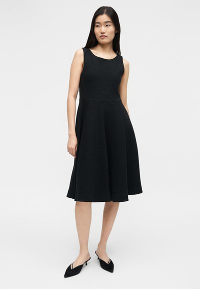 Γυναίκα που φοράει αμάνικο, μαύρο φόρεμα μήκους μέχρι το γόνατο με εφαρμοστή μπουστά και φουσκωτή φούστα, συνδυασμένο με μαύρα πέδιλα στιλέτο με ανοιχτή πτέρνα.