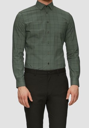 Mand iført en tætsiddende grøn langærmet skjorte med sorte tern og sorte bukser, stående mod en ensfarvet baggrund.