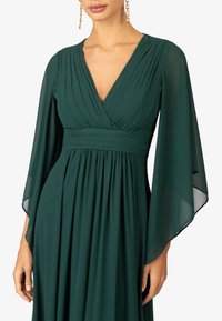 Abito verde smeraldo con scollo a V profondo, corpino plissettato e maniche a campana fluide realizzate in tessuto leggero. Eleganti drappeggi sotto la vita.