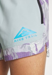 Shorts gris claro con un patrón de camuflaje púrpura, con detalles en negro, un bolsillo lateral y un logotipo de Nike Trail en azul.