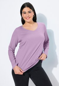 Fialový dlouhý rukáv s výstřihem do V, vyrobený z měkké tkaniny, s uvolněným střihem a hladkou texturou. V páru s tmavými úzkými džínami.