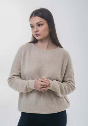 Beżowy, miękki sweter z okrągłym dekoltem i długimi rękawami, o luźnym kroju i ściągaczach przy mankietach, na jasnym tle.