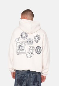 Cremfarvet hættebluse med et stort grafisk print af forskellige frimærker og designs i sort på ryggen. Har snore i hætten og en afslappet pasform.