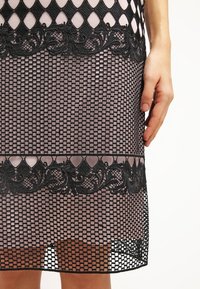 Czarna koronkowa sukienka z diamentowym wzorem siateczkowym, z falbanką z koronki u dołu i misternymi haftami. Podstawa jest w delikatnym różowym odcieniu.