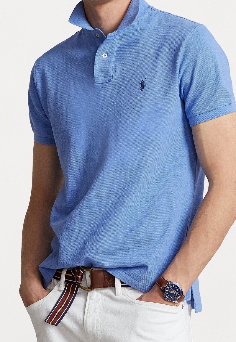 Homme portant un polo bleu clair avec un logo foncé, un pantalon blanc, une ceinture rayée et une montre au cadran bleu avec un bracelet en cuir marron.