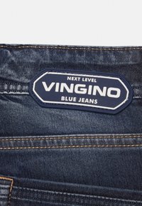 Blåjeans med en marinblå etikett med vit текст som läser "NEXT LEVEL VINGINO BLUE JEANS", accentuerade med ljusbruna sömmar.