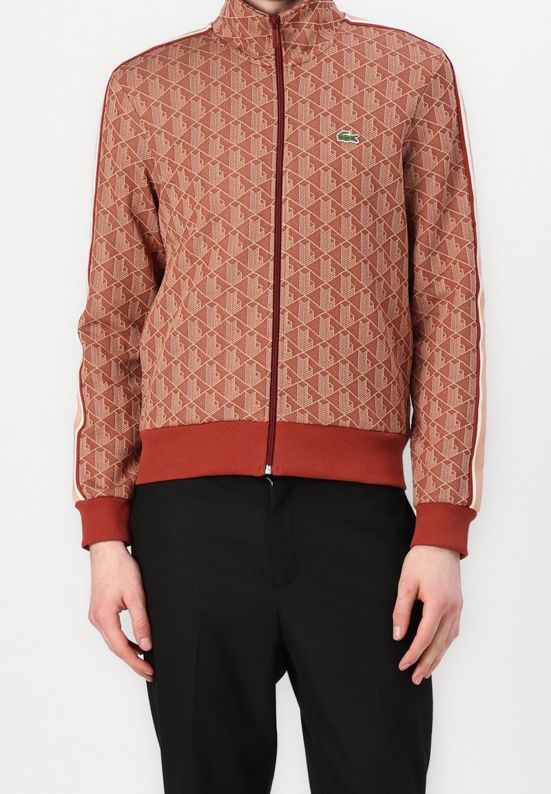 Homme portant une veste zippée rouge rouille avec un motif géométrique, des poignets et un ourlet côtelés, des bandes latérales, et un petit logo crocodile vert sur la poitrine.