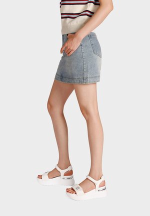 Donna che indossa una minigonna di denim chiaro, top senza maniche a righe e sandali bianchi con zeppa, in piedi davanti a uno sfondo semplice.