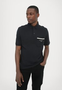 Svart polo-skjorte med to-knaps lukking og korte ermer. Har lomme med hvit stripe og merke tekst, laget av et strukturert stoff.