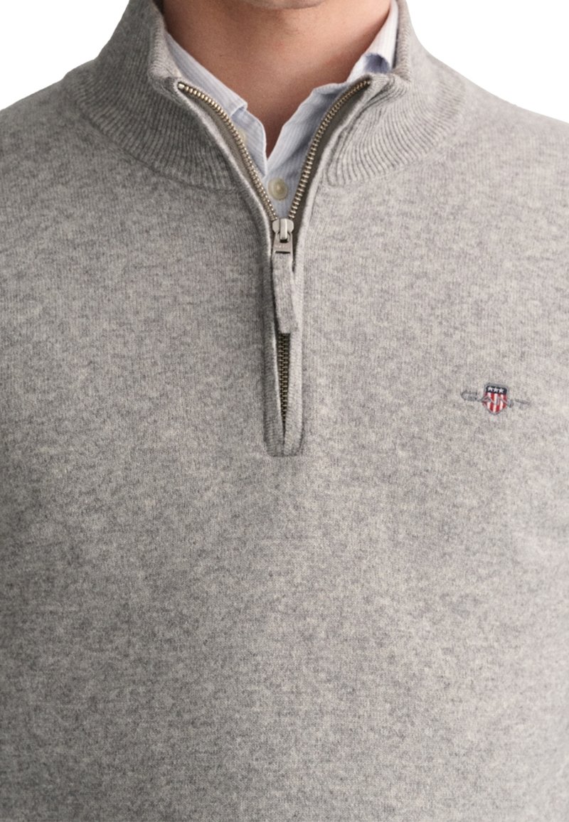 Homme portant un pull léger gris clair à fermeture éclair par-dessus une chemise à col bleu clair avec un petit blason brodé sur la poitrine.