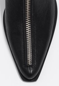 Bota de cuero negra de tobillo con cierre de cremallera plateada, superficie texturizada y suela plana elegante. Presenta un diseño minimalista.