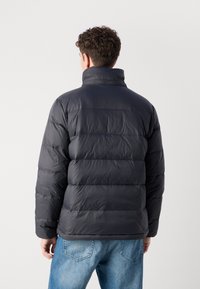 Donkere puffy jas met een hoge kraag, voorzien van horizontale stiksels en elastische manchetten, gecombineerd met lichtblauwe jeans.