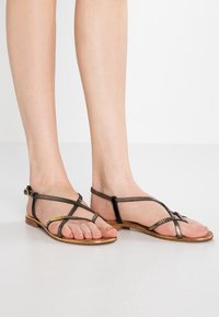 Des sandales plates en bronze métallique présentent de fines lanières avec un design tressé et une petite boucle, posées sur une surface blanche lisse.