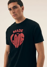 Czarna bawełniana koszulka z krótkim rękawem, ozdobiona dużym, czerwonym sercem z napisem "STWORZONA DO MIŁOŚCI" w pogrubionej, stylizowanej czcionce. Okrągły dekolt.