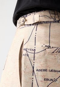 Beige trousers with a map print, featuring a silver buckle belt, side pocket, and stitched detail along the waistband.