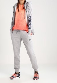 Sweat à capuche gris zippé avec logo sur la manche, tee-shirt de sport orange, pantalon de survêtement gris avec logo, et baskets noires avec accents roses.