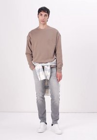 Sudadera de manga larga color topo con un pequeño logo, combinada con jeans grises claros y zapatillas blancas. Una camisa de cuadros está atada a la cintura.
