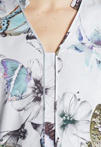 Blusa floreale con scollo a V, decorata con farfalle e fiori colorati in blu, verde e nero su uno sfondo bianco. Tessuto morbido.