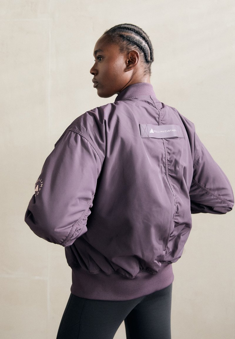 Lila bomberdzseki magas gallérral, rugalmas mandzsettával és aljjal. Hátán logóval és a bal ujján rózsaszín részlettel rendelkezik. Simább textúrájú.