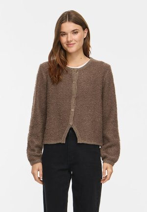 Junge Frau mit braunen Haaren, die einen braunen, flauschigen, geknöpften Cardigan und schwarze Hosen trägt und vor einem weißen Hintergrund steht.