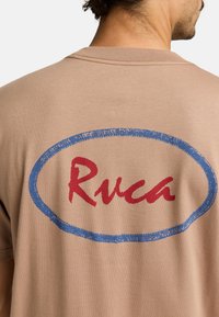 Béžové bavlněné tričko s velkým modrým oválným obrysem a červeným ručně psaným textem "Ruca" na zádech. Krátké rukávy a kulatý výstřih.