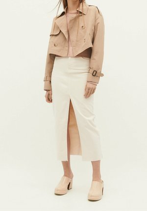Femme portant un trench beige court, un top rose, une jupe blanche taille haute avec une fente devant, et des mules à talons hauts beiges, debout sur un fond uni.