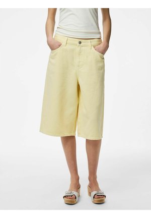 Persoană purtând pantaloni scurți palid galbeni, lungime până la genunchi, cu picioare largi, bluză albă fără mâneci și sandale bej cu platformă și catarame. Mâinile în buzunare.