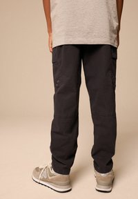 Schwarze Cargo-Hosen mit seitlichen Taschen, elastischen Bündchen und Metallösen. Kombiniert mit grauen Wildleder-Sneakers, die ein leichtes Mesh-Detail zeigen.