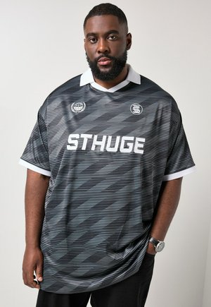 Mężczyzna z brodą ubrany w czarno-szarą sportową koszulkę w paski z białym kołnierzem i napisem „STHUGE”, stojący na jednolitym tle.