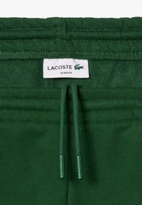 Pantalons de survêtement slim verts avec une taille côtelée, un cordon de serrage et une étiquette avec le logo Lacoste. Texture douce, couleur uniforme, design minimaliste.